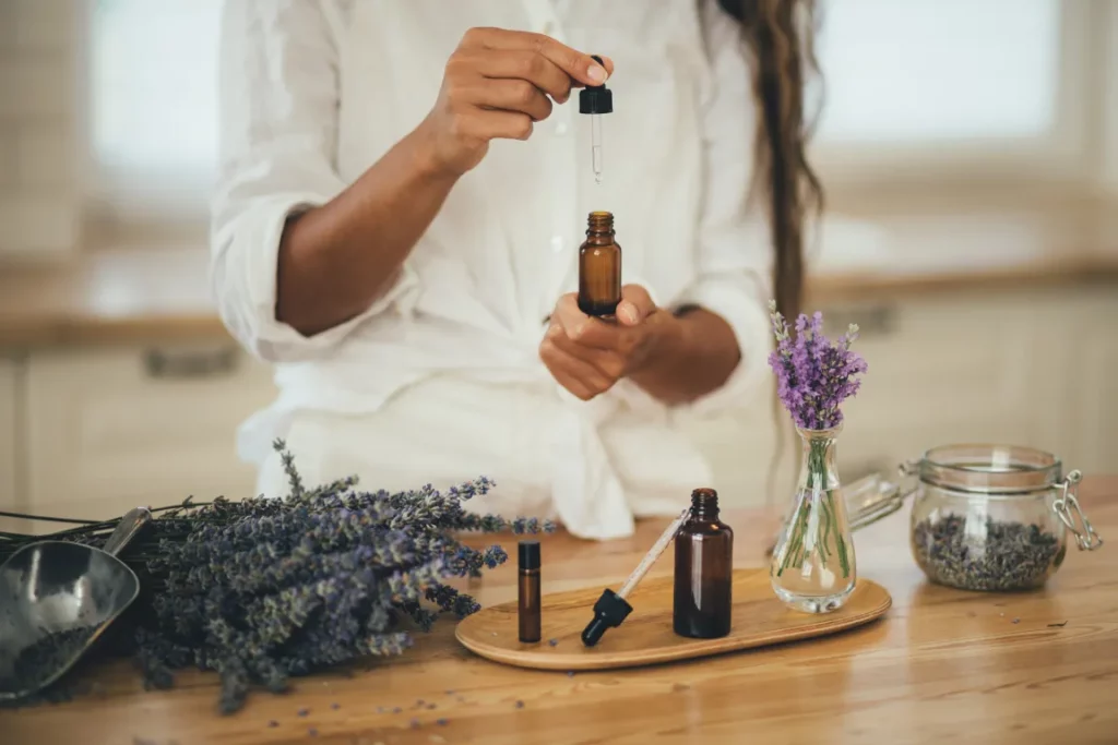 An aromatherapist wearing a white shirt and pants holds an essential oil bottle and pipette in front of a table with a shovel, dried lavender, essential oil bottles, and a container of lavender petals.